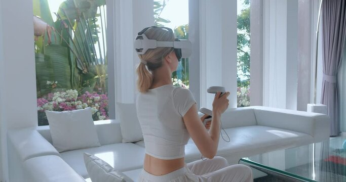Female playing games on virtual reality device. Woman using virtual reality headset. Female gamer uses virtual reality headset and joystick controllers for active game playing, sitting on sofa at home