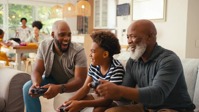 Multi-generation male family sitting on sofa at home holding controllers playing video game together with father cheating and covering son's eyes - shot in slow motion