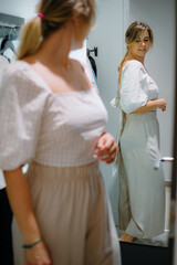 Fototapeta premium Glorious lady in white blouse and beige trousers turning sideways and smiling standing in front of mirror in wardrobe.