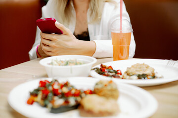 Young woman freelancer sitting at table with dishes in cafe and holding phone. Concentrated lady working at lunch time.