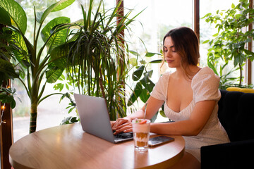 Young beautiful woman wearing dress sitting at table with laptop working remotely on new web design project in summer cafe with green plants