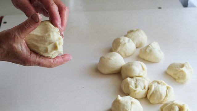 Close-up of wrinkled hands of an elderly woman pinching off raw dough to form balls. An old grandmother bakes buns using a traditional family recipe.