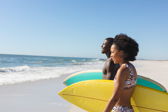 Happy, fit african american couple carrying surfboards walking on sunny beach to the sea, copy space