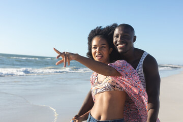 Happy african american couple embracing and pointing away on sunny beach by sea