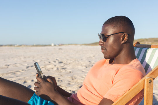 Happy african american man in sunglasses sitting in deckchair using smartphone on sunny beach