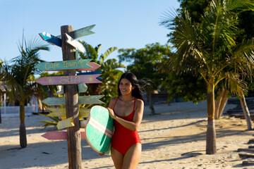 Happy biracial woman in red swimsuit carrying surfboard on sunny beach