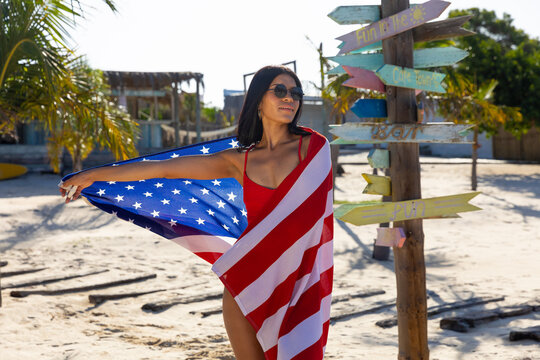 Happy biracial woman in sunglasses and red swimsuit dancing with american flag on sunny beach - Powered by Adobe