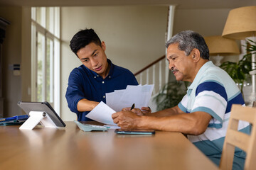 Diverse financial advisor and senior man discussing paperwork and using tablet in dining room