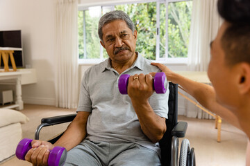 Diverse male physiotherapist advising and senior male patient in wheelchair using dumbbells