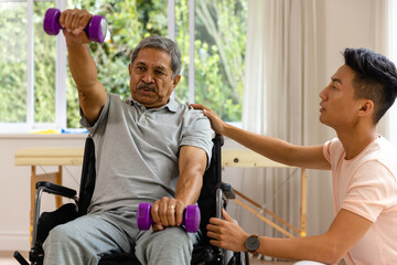 Diverse male physiotherapist advising and senior male patient in wheelchair using dumbbells