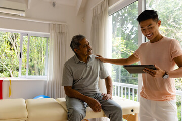 Happy diverse male physiotherapist examining senior male patient using tablet