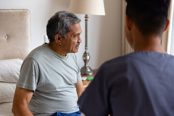 Diverse male doctor and senior male patient sitting on bed and discussing at home