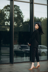 A young woman in a black suit drinks coffee and poses on the street with a laptop. Business portrait