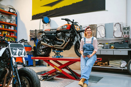 Smiling mechanic woman holding digital tablet sitting over platform with custom motorcycle on factory