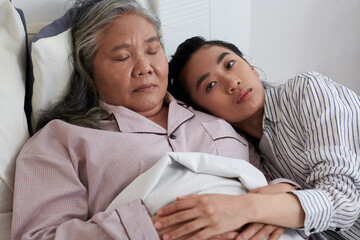Pensive serious young woman leaning on shoulder of sick sleeping mother