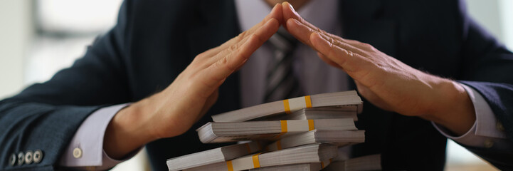 Banker making roof with hands over bundles of dollar bills closeup. Safe storage of money and profitable financial investments concept
