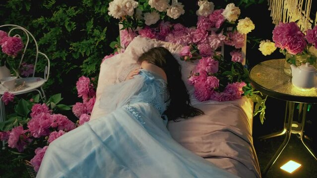 Fairy image sleeping beauty. Fantasy woman sleep on medieval bed. Long loose hair lies on soft pillow. blooming pink white peonies flowers. Spring green foliage tree. Blue vintage dress back rear view