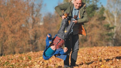 A father spins his son by his feet in the park in the fall.