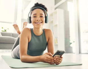 Tuinposter Yoga, portrait and happy woman relax on a living room floor with music, headphones and phone for streaming at home. Face, smile and lady on fitness break with social media, radio or podcast in lounge © HockleyMedia/peopleimages.com