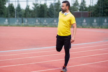 Obraz premium Young man with wireless headphones warming up leg muscles before training on running track with rubber surface and white lines at sports ground in city park