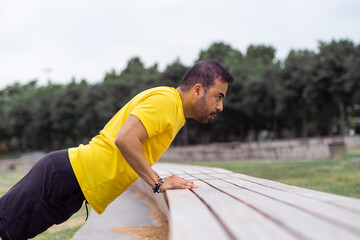 Bearded sportsman doing push-ups leaning on long bench in summer city park young athlete practicing exercises for arms muscles growing on sports arena