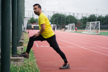 Obraz premium Athletic man in tracksuit doing lunges stretching legs leaning on fence support at urban stadium runner warming up training on sports arena with football field