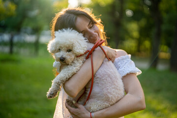 Content young dark haired woman in casual wear holding in arms an adorable Bichon Frise dog while resting in park during summer daytime