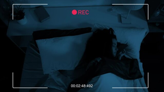 Security camera shot of young woman wearing nightgown turning off bedside lamp and going to sleep in her bed