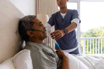 Diverse male doctor giving oxygen mask to senior male patient in bed at home