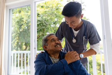 Happy diverse male doctor discussing with senior male patient in wheelchair at home