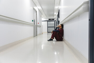 Tired caucasian female doctor wearing scrubs, sitting on floor in corridor at hospital, copy space