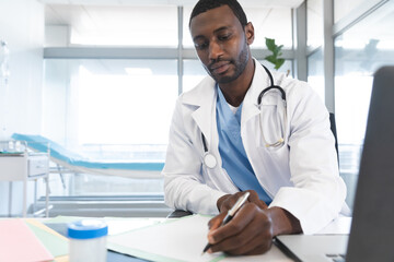 Naklejka na ściany i meble African american male doctor wearing lab coat and stethoscope, sitting at desk, taking notes