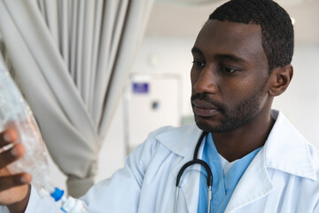 African american male doctor wearing lab coat, applying drip in hospital room