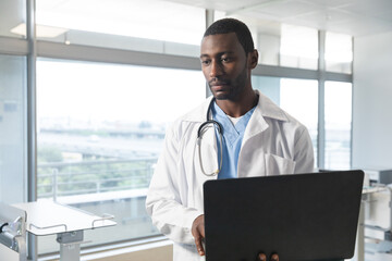 Fototapeta premium African american male doctor wearing lab coat and stethoscope, using laptop at hospital