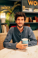 Delighted bearded male with curly hair, sitting at table in cafe and enjoying weekend while looking at camera.