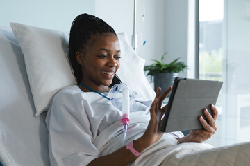 Happy african american female patient lying on bed and using tablet in hospital room