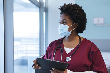 African american female doctor wearing scrubs and face mask, looking through window