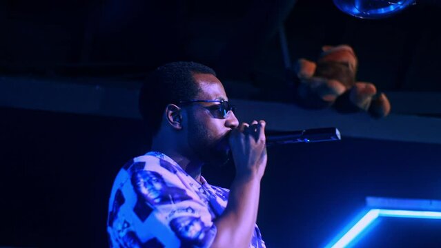 Black african male man artist singer rapper showman performs concert party on the club stage in dark scenery with neon lights blue theme and makes speech