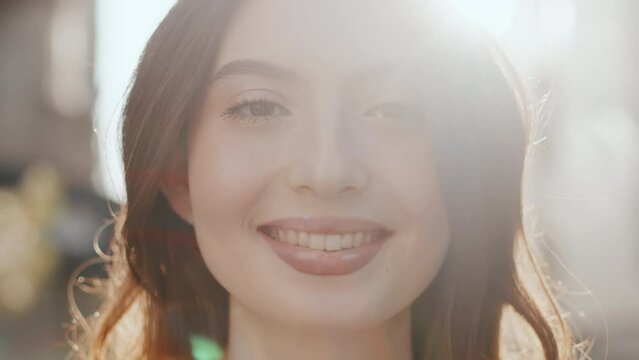 Portrait of an attractive young woman close up who smiling outdoors while looking at the camera in a counter sunlight. Concept of urban carefree summer life