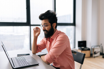 Side view of confident Indian business man in eyeglasses talking online by video call on laptop sitting at desk on background of window. Stylish freelancer male using computer for video conversation.