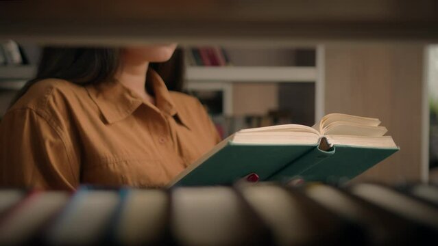 Unrecognizable woman reading book in university library near bookcase smart female girl college student teacher read textbook literature prepare for exam studying homework view through bookshelf books