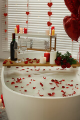 Wooden tray with wine, burning candles and rose petals on tub in bathroom. Valentine's day celebration