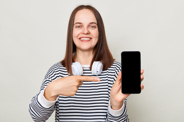 Beautiful smiling happy cheerful brown haired adult woman wearing striped shirt standing isolated over gray background pointing at smart phone empty display.