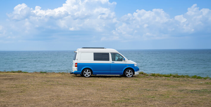 Blue vintage camper van parked on a grassy plain with the Atlantic Ocean in the background under a semi-cloudy sky lit by the early morning sun.
