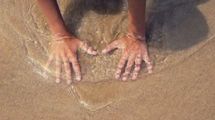 The hands of a teenager lead along the sand and small waves cover them from above. body part in...