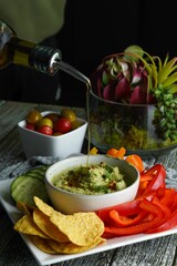 An olive oil being poured on a dipping sauce surrounded by chips and vegetables