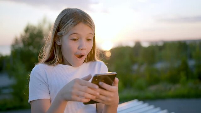 Little smiling cute girl using smartphone at sunset, typing message. Emotions good news surprise concept. Kid girl holding smart phone enjoying using technology device, mobile apps, playing games