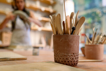 Cup with set of dirty art and craft sculpting tools on wooden table in pottery workshop, used to work clay and ceramic
