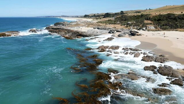 Aerial of the foamy sea waves hitting the rocks and the sandy shore in Otago, New Zealand