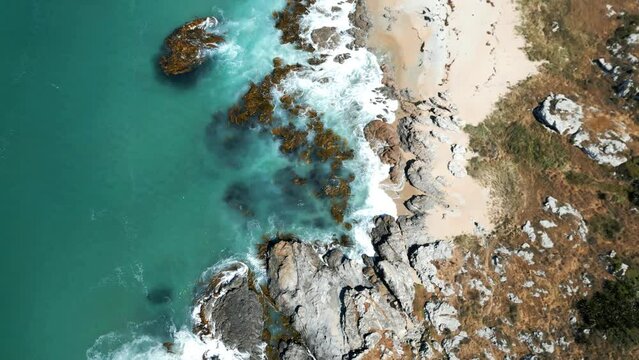 Top view of the foamy sea waves hitting the rocks and the sandy shore in Otago, New Zealand